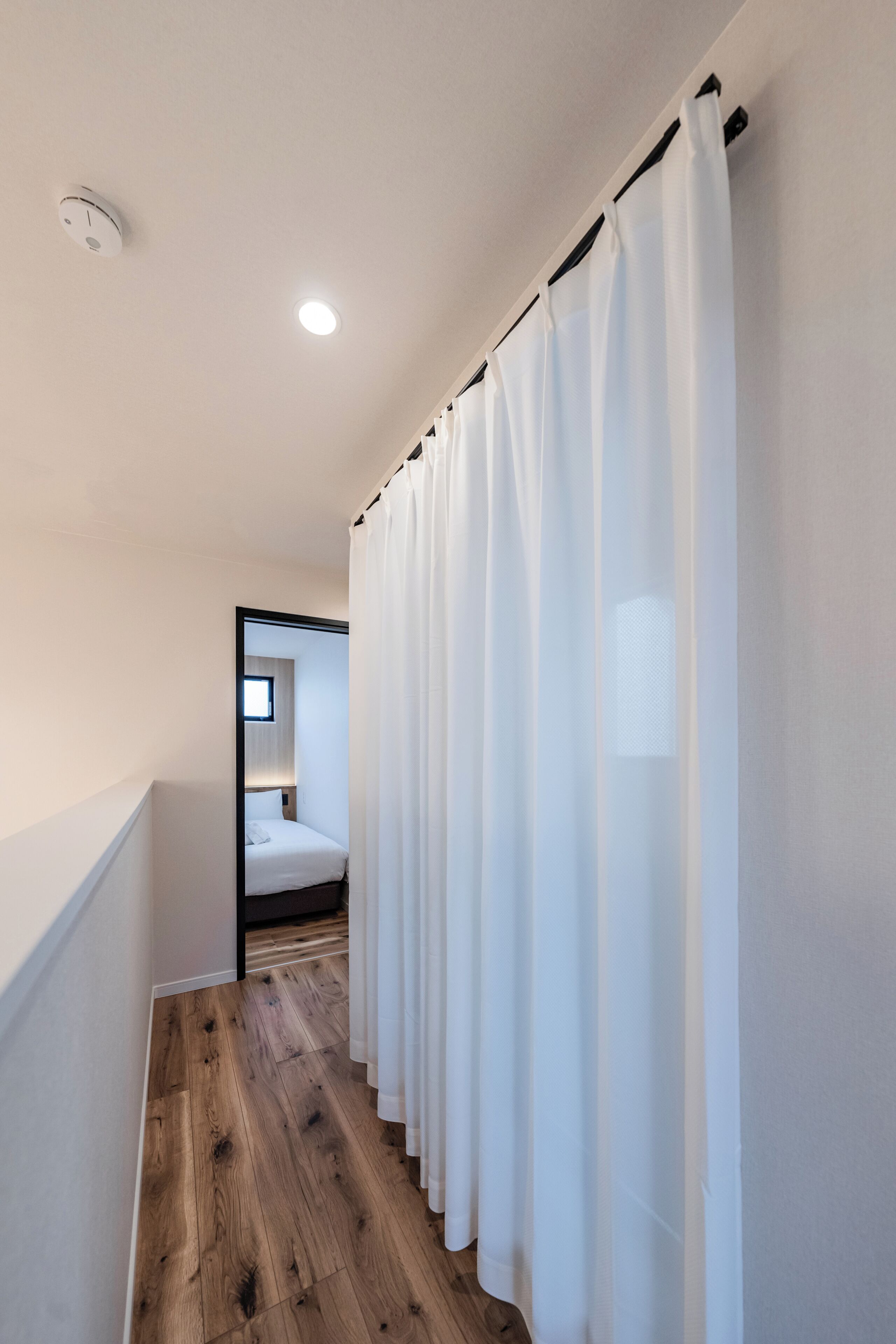 A narrow hallway with wooden flooring, white walls, and a white curtain. A small window with a view of a bedroom is visible in the distance.