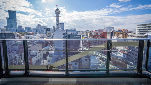 Vista de un paisaje urbano con una torre destacada y varios edificios, tomada desde un balcón con barandilla de vidrio.