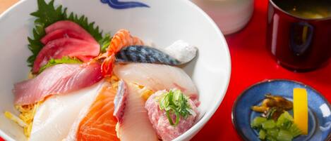 Un bol de sashimi composé de différents types de poisson et agrémenté de garnitures, une petite portion de wasabi et une tasse de thé, le tout posé sur une table rouge.