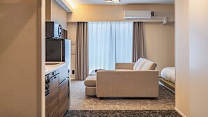 Una habitación de hotel compacta con kitchenette, un sofá, una cama y un ventanal grande con cortinas.
