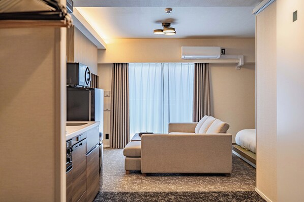 A compact hotel room with a kitchenette, a sofa, a bed, and a large window with curtains.