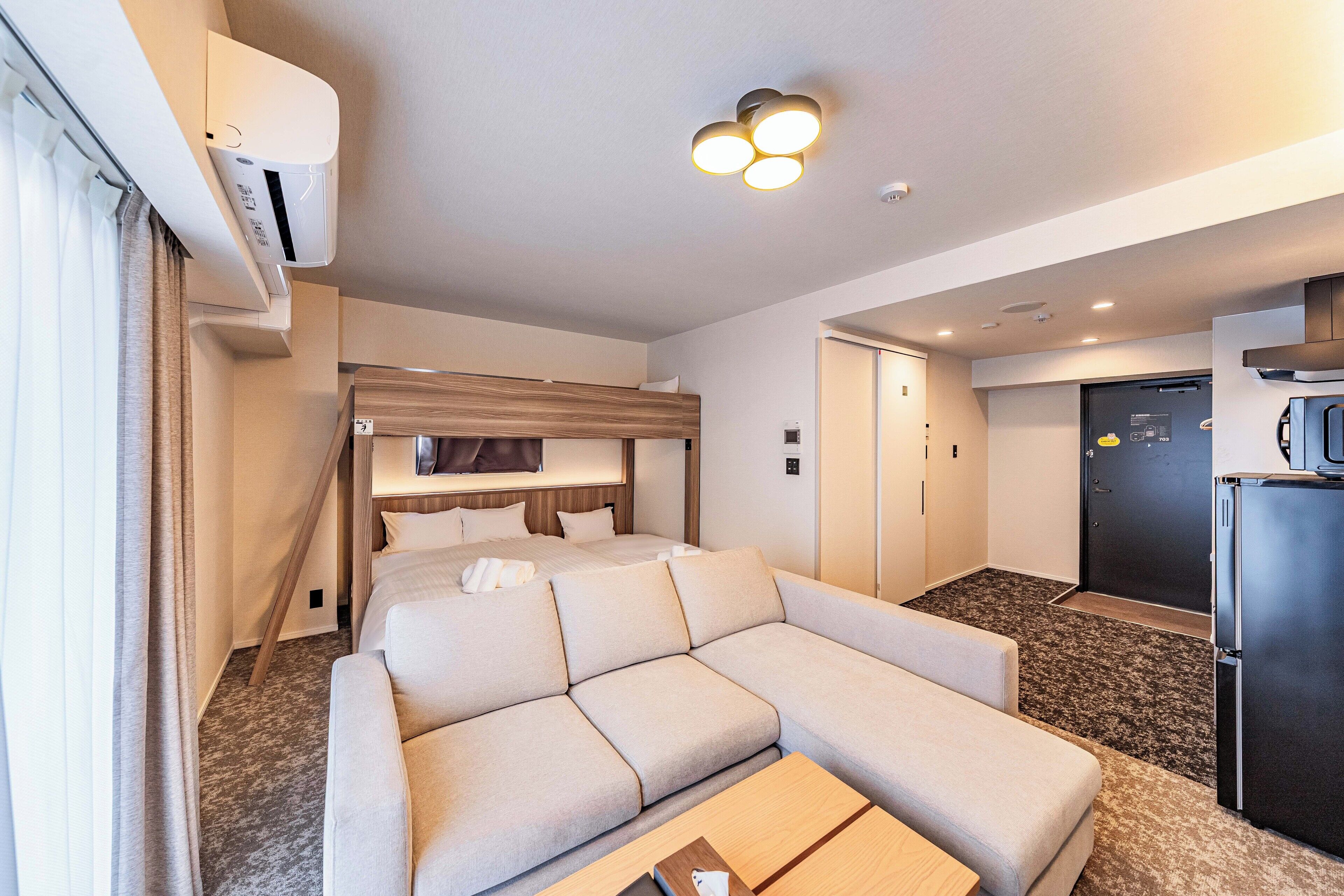 Una habitación de hotel moderna con sofá cama, una mesita de madera, un banco integrado, refrigerador y una pequeña zona de cocina.