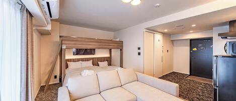 Una habitación de hotel moderna con sofá cama, una mesita de madera, un banco integrado, refrigerador y una pequeña zona de cocina.