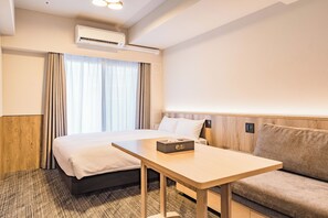 A hotel room with a bed, a bedside table, a sofa, a window with curtains, an air conditioning unit, and a wall-mounted control panel.