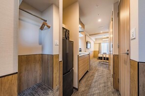 Pasillo de hotel con suelo de madera, un refrigerador, un lavamanos y una cama a lo lejos.