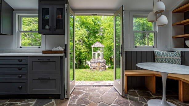 Una cocina con armarios grises, piso de piedra y vista a un jardín a través de una puerta abierta.