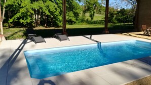 Une piscine rectangulaire, bordée d’une terrasse carrelée, entourée d’arbres et d’une pelouse.