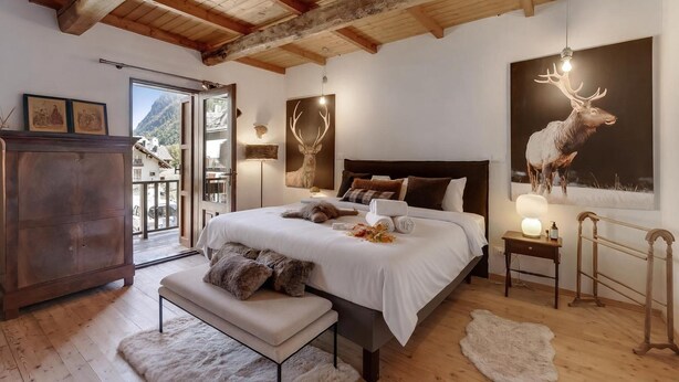 A bedroom with a large bed, wooden ceiling beams, a view of mountains, a deer wall art, and a rustic wooden cabinet. - La Baita 1800 Ski In Ski Out (Claviere)