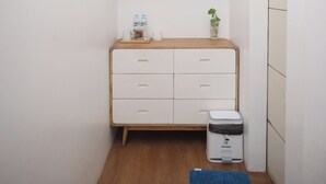 Ett litet, rent rum med trägolv, en vit byrå med lådor, en liten papperskorg, en blå matta och en luftkonditioneringsenhet på väggen.