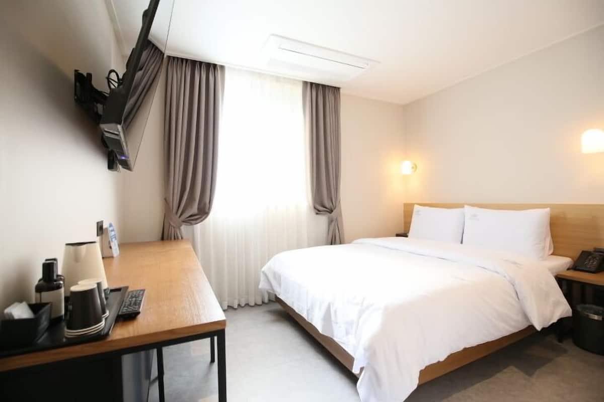 A hotel room with a bed, a desk, a lamp, a phone, and a window with curtains.