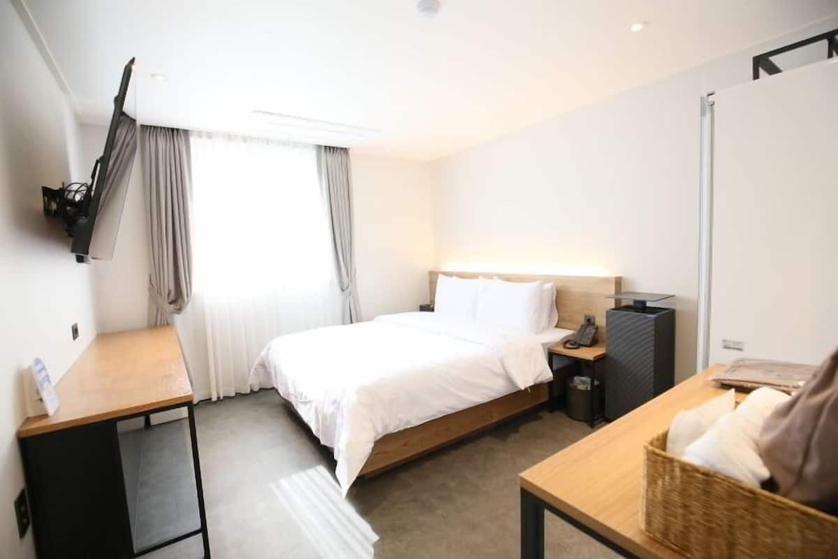 A hotel room with a bed, a desk, a chair, a TV mounted on the wall, and a window with curtains.