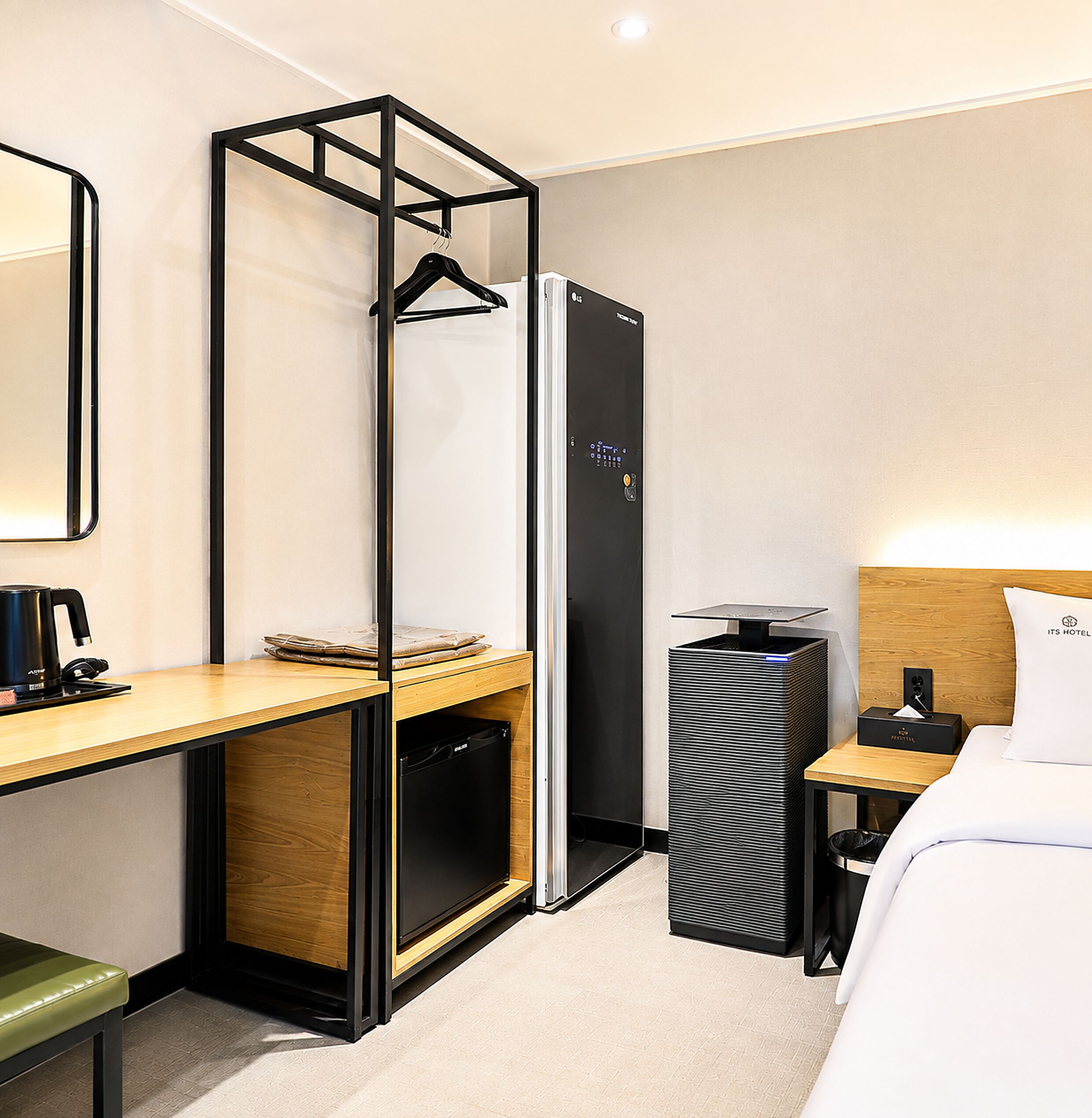 A hotel room with a bed, a desk with a chair, a small refrigerator, a trash bin, and a bedside table with a lamp.