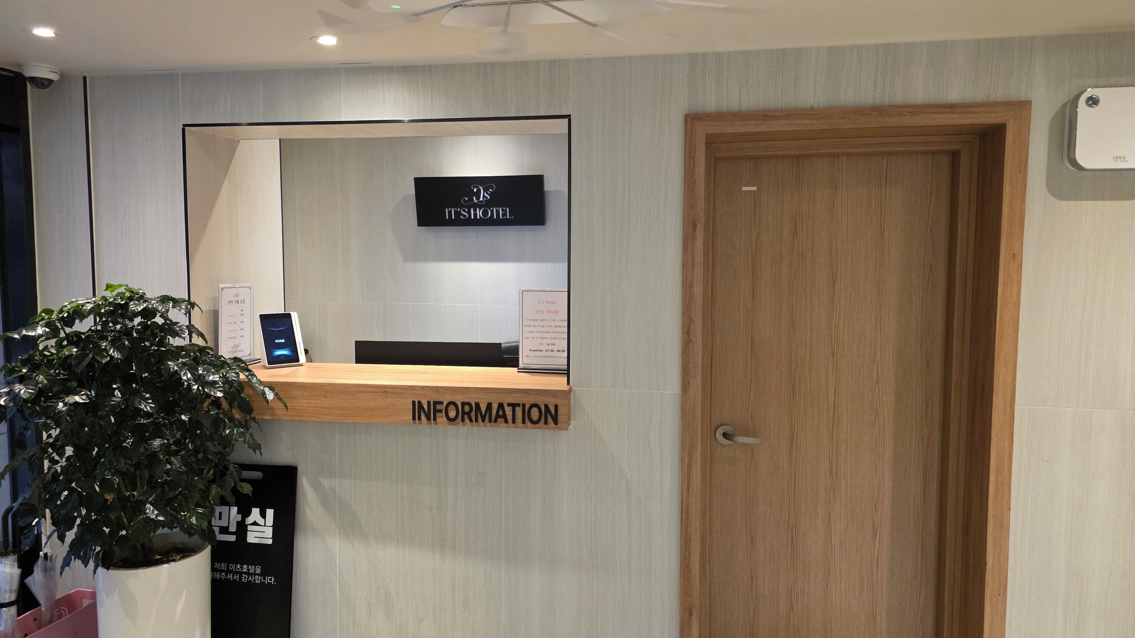 A reception desk with a digital screen, a wooden door, and a sign indicating hotel information.