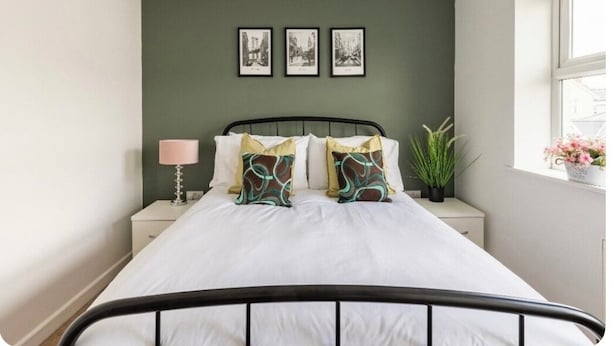 A bedroom with a bed, bedside tables, a lamp, framed pictures on the wall, and a window with a plant. - Town center white house (Bournemouth)