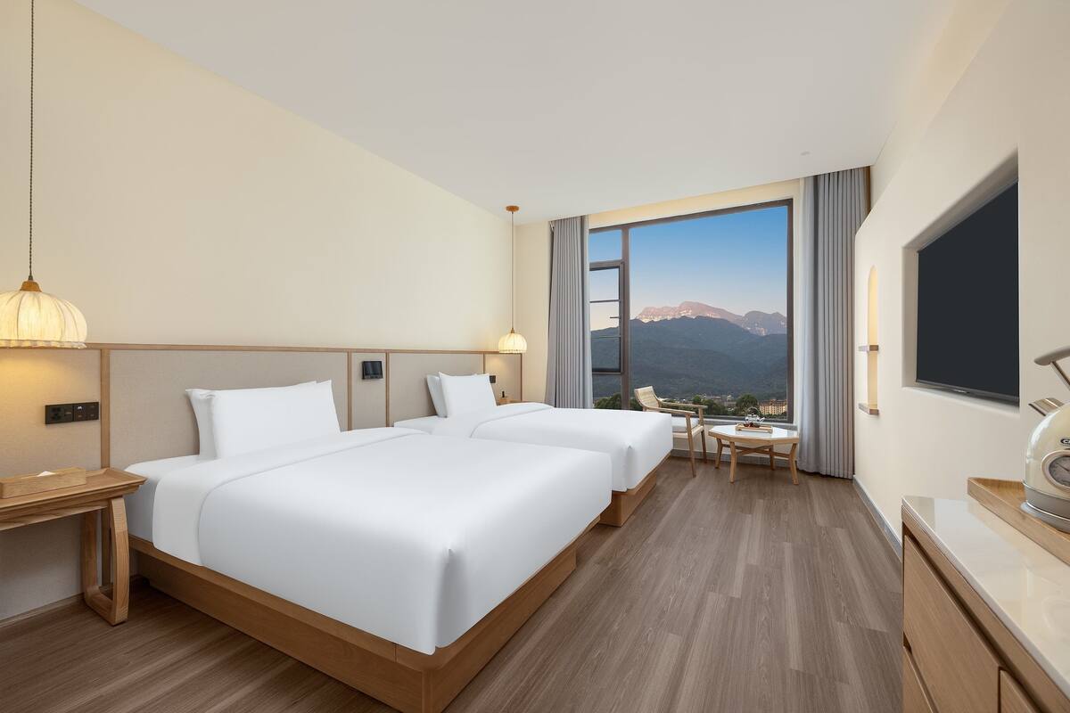 A hotel room with two beds, a desk, a chair, a TV, and a large window with a mountain view.