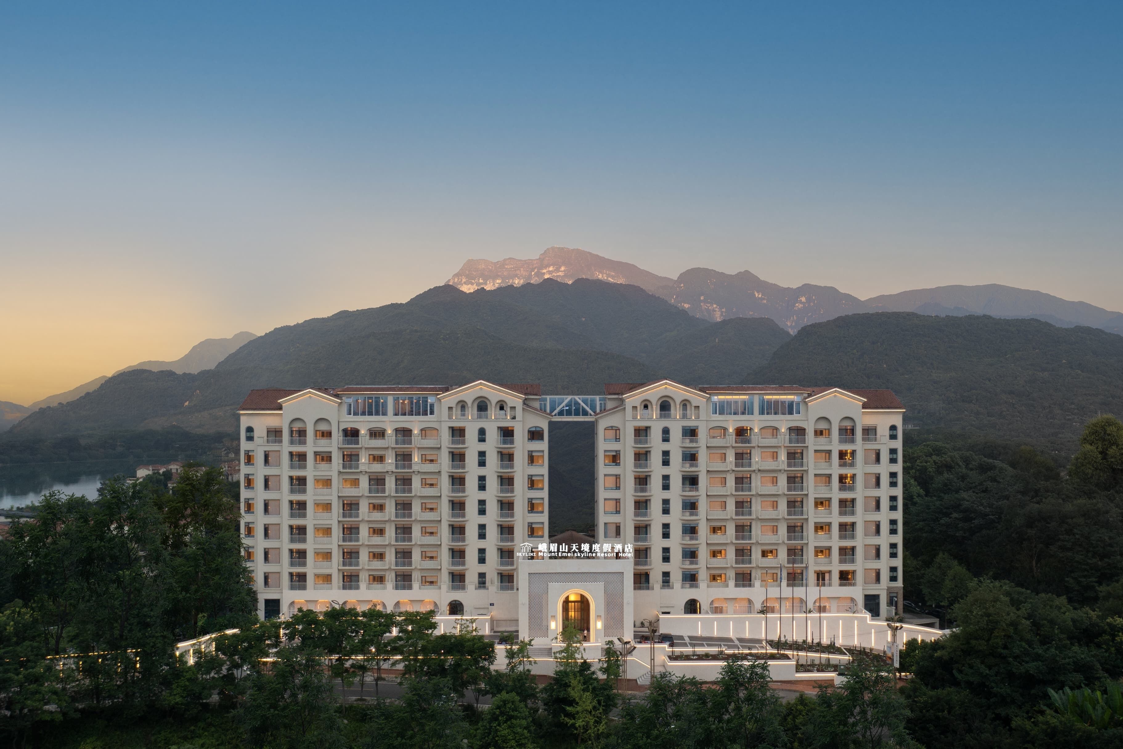 A large hotel with multiple floors and a central entrance, surrounded by lush greenery and mountains in the background.