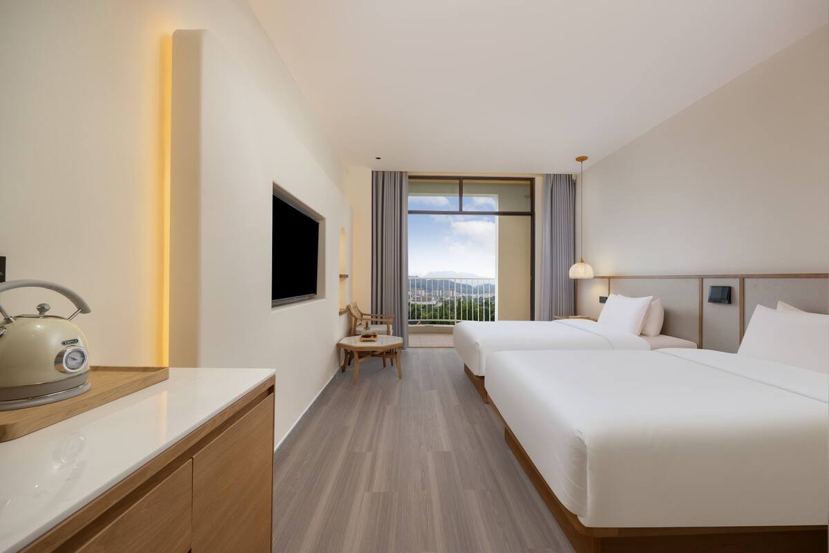 A hotel room with two beds, a TV, a kettle, a small table, and a balcony with a view.