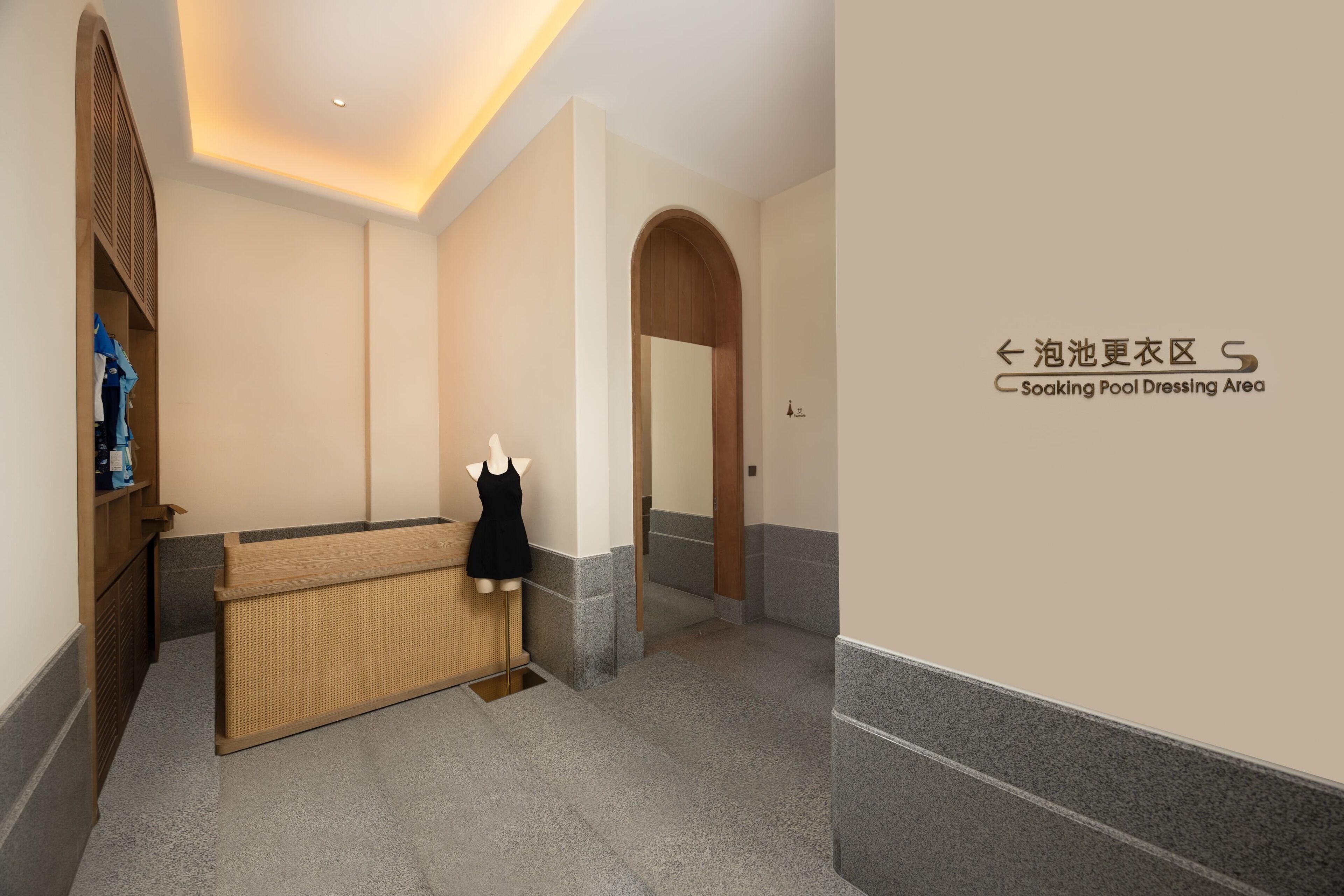 Zona de vestuarios de piscina con mostrador de recepción de madera, un maniquí vestido de negro y suelo de baldosas.