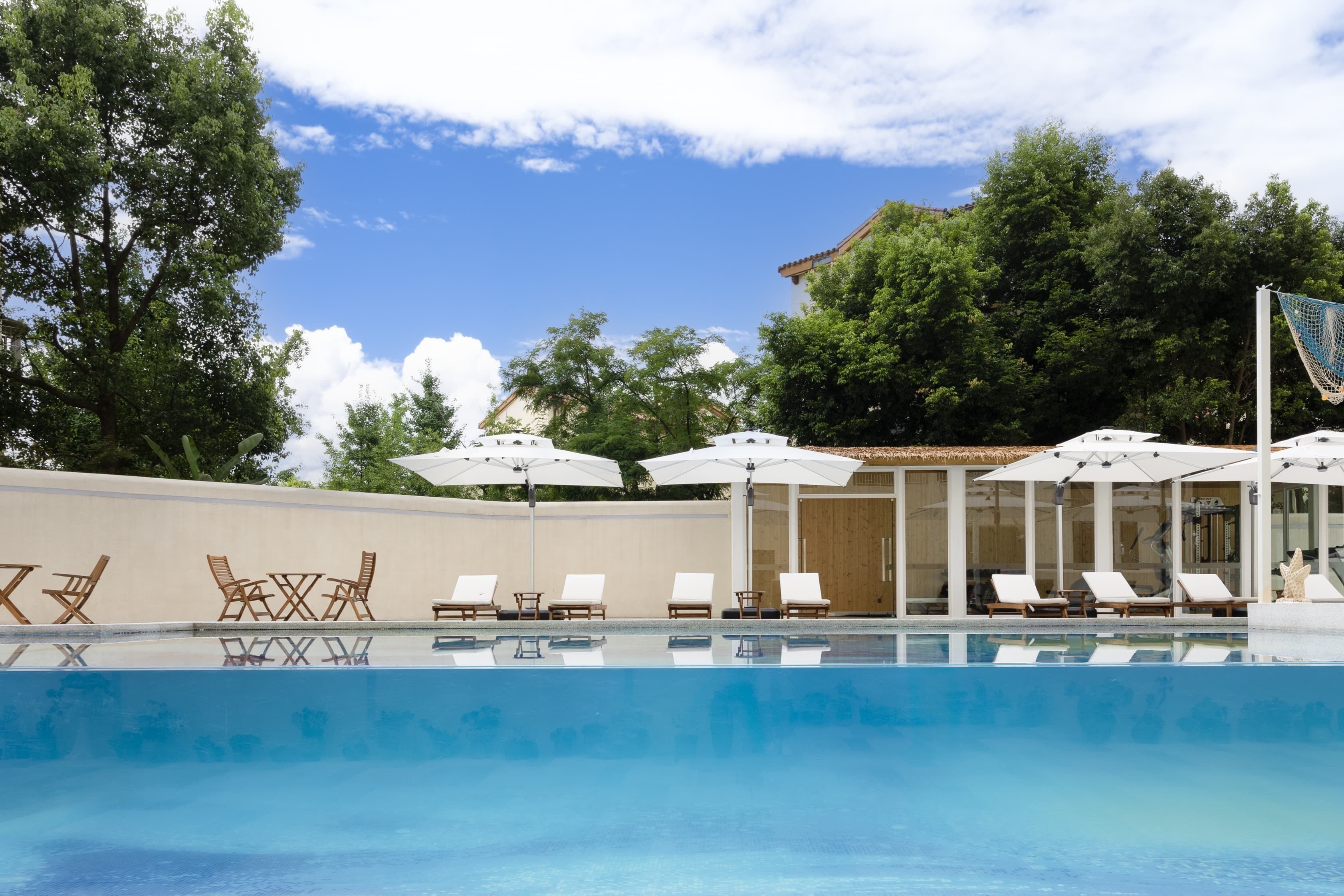 A swimming pool with lounge chairs and umbrellas, surrounded by trees and a building.