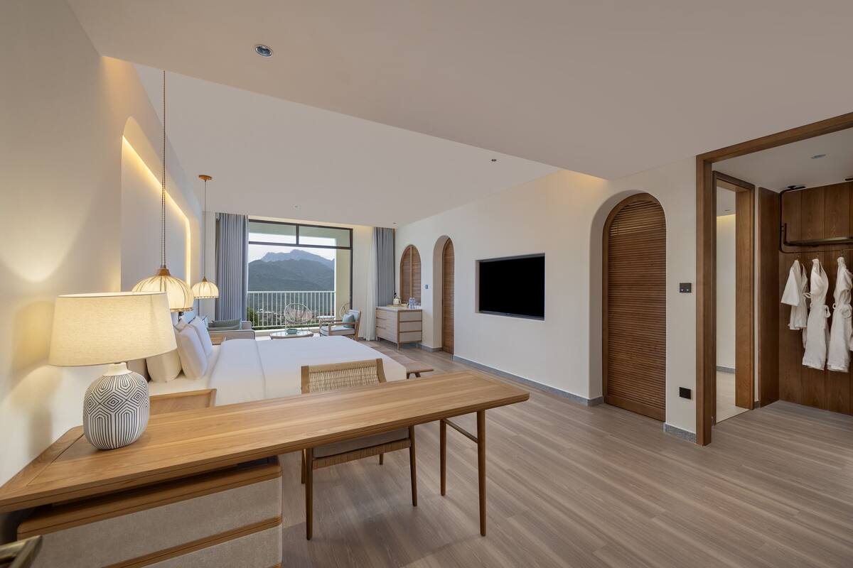 A modern hotel room with a wooden dining table, a sofa, a TV mounted on the wall, and a view of mountains through the window.