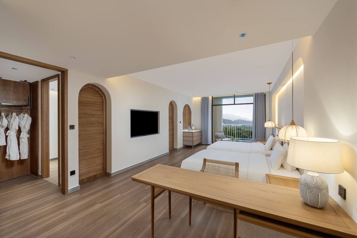 A modern hotel room with a wooden floor, a bed with white bedding, a desk with a lamp, a TV, and a view of the outdoors.