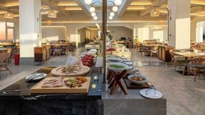 Ένας ευρύχωρος χώρος εστίασης στο ξενοδοχείο, με ψηλό ταβάνι και φεγγίτη, που διαθέτει πολλά τραπέζια και μια κεντρική προθήκη με διάφορα είδη φαγητού σε στυλ μπουφέ.