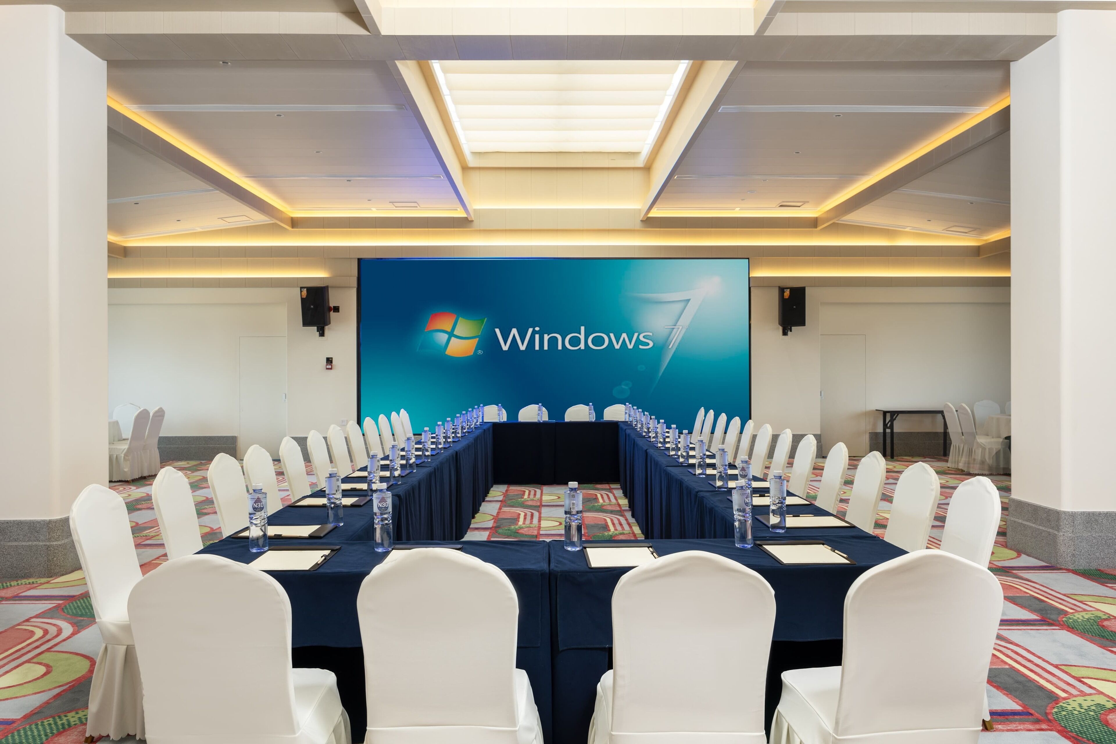 Un salón de conferencias con una larga mesa, sillas blancas y una pantalla grande mostrando el logotipo de Windows 7.