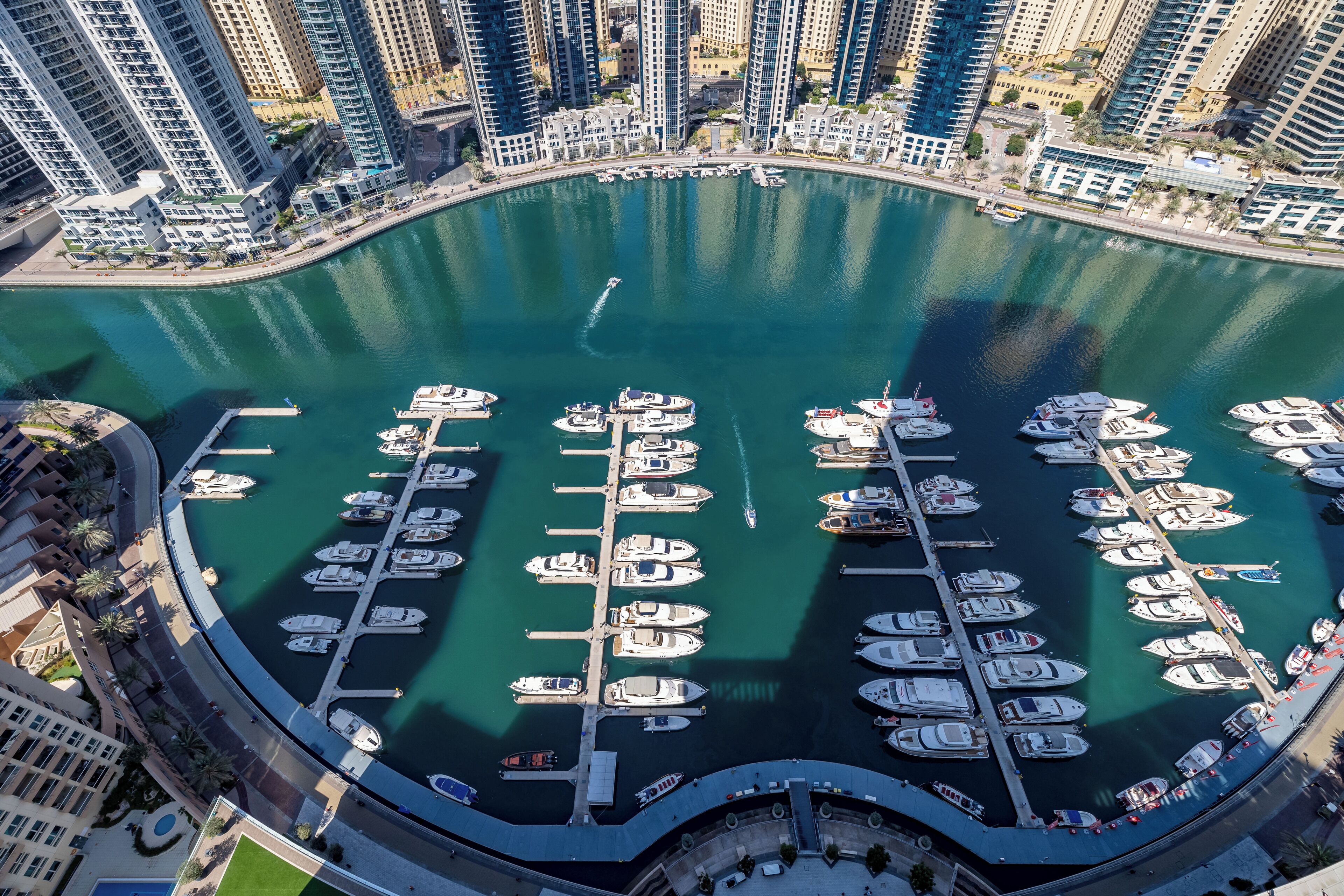 An aerial view of a marina with numerous boats docked in a circular layout, surrounded by high-rise buildings.