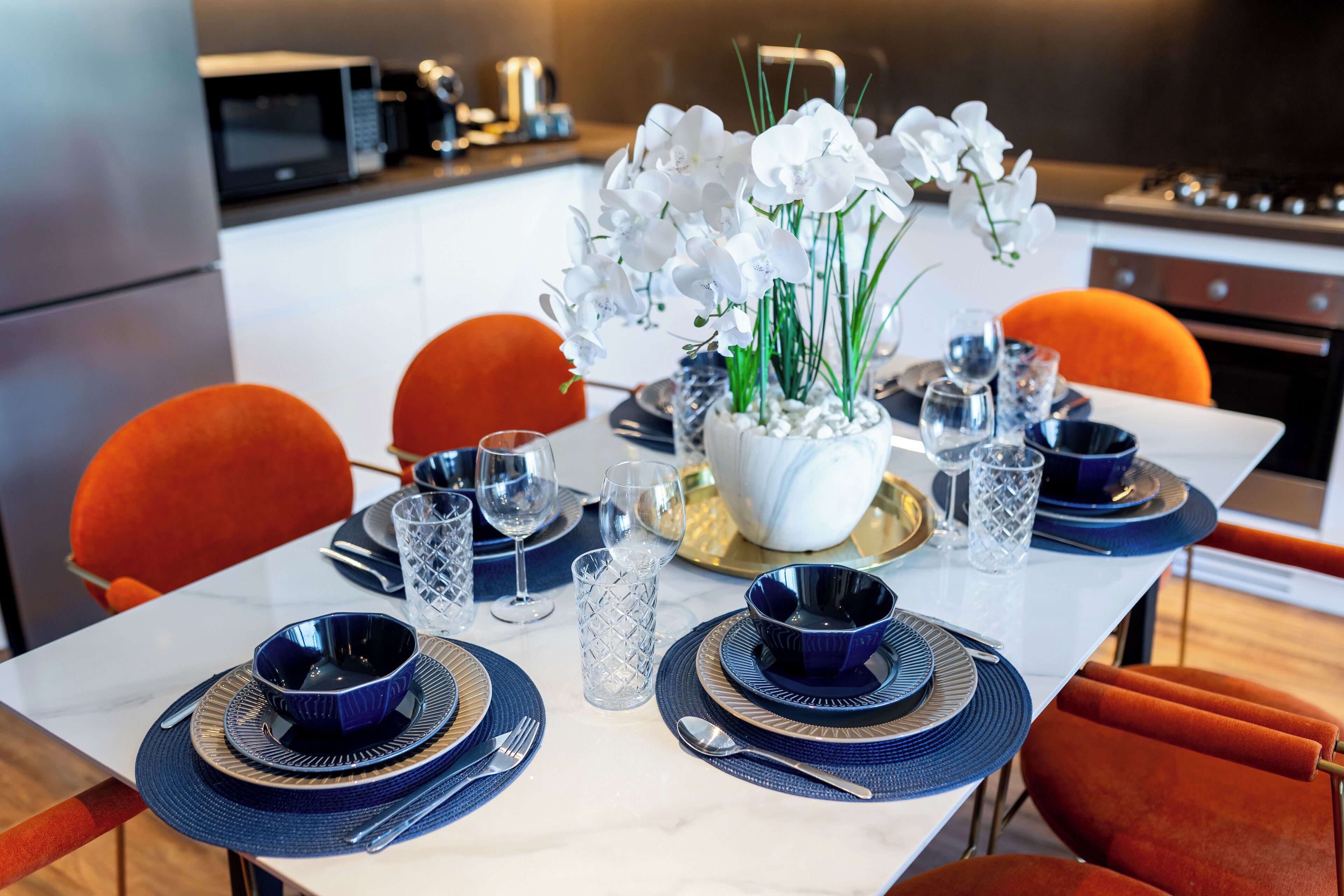 A modern dining area with a white table set for four, featuring blue and gold accents, white orchids, and orange chairs.