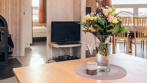 Ein modernes Wohnzimmer mit Holzboden, einem Flachbildfernseher auf einem Schrank, einem Essbereich mit Holzstühlen und einem Blumenstrauß auf einem Tisch.