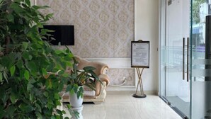 A modern lobby with a beige sofa, a potted plant, a wall-mounted TV, and a glass door leading outside. - White Hotel (Thanh Vinh)