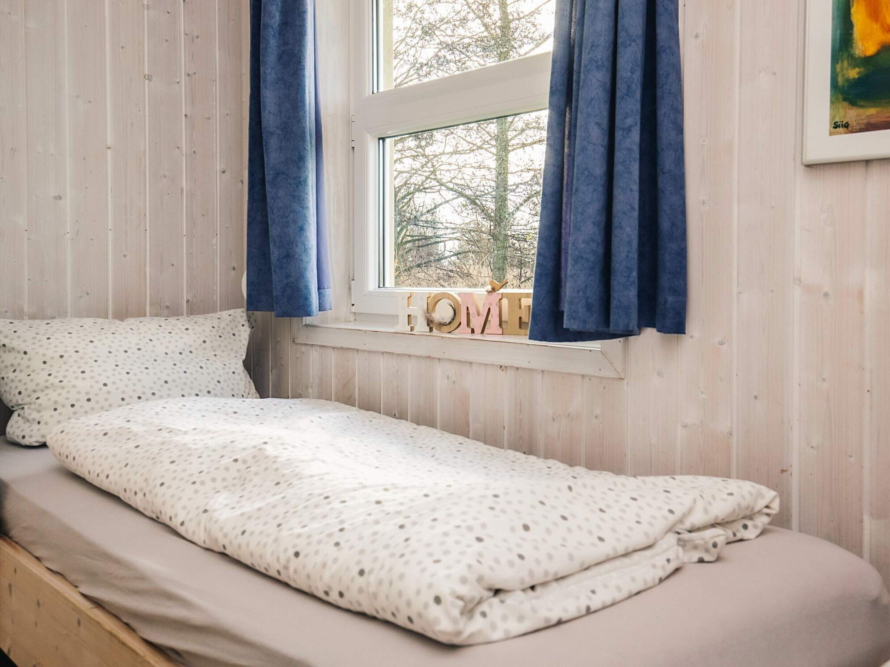 A small bed with a polka dot bedspread, a window with blue curtains, and a 'HOME' sign on the sill.
