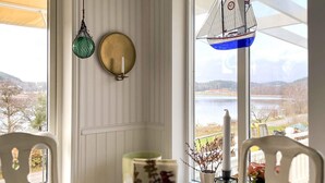 A dining area with a table set for two, a hanging sailboat decoration, and a view of a lake and hills through large windows. - 5 Star Holiday Home in Uddevalla (Uddevalla)
