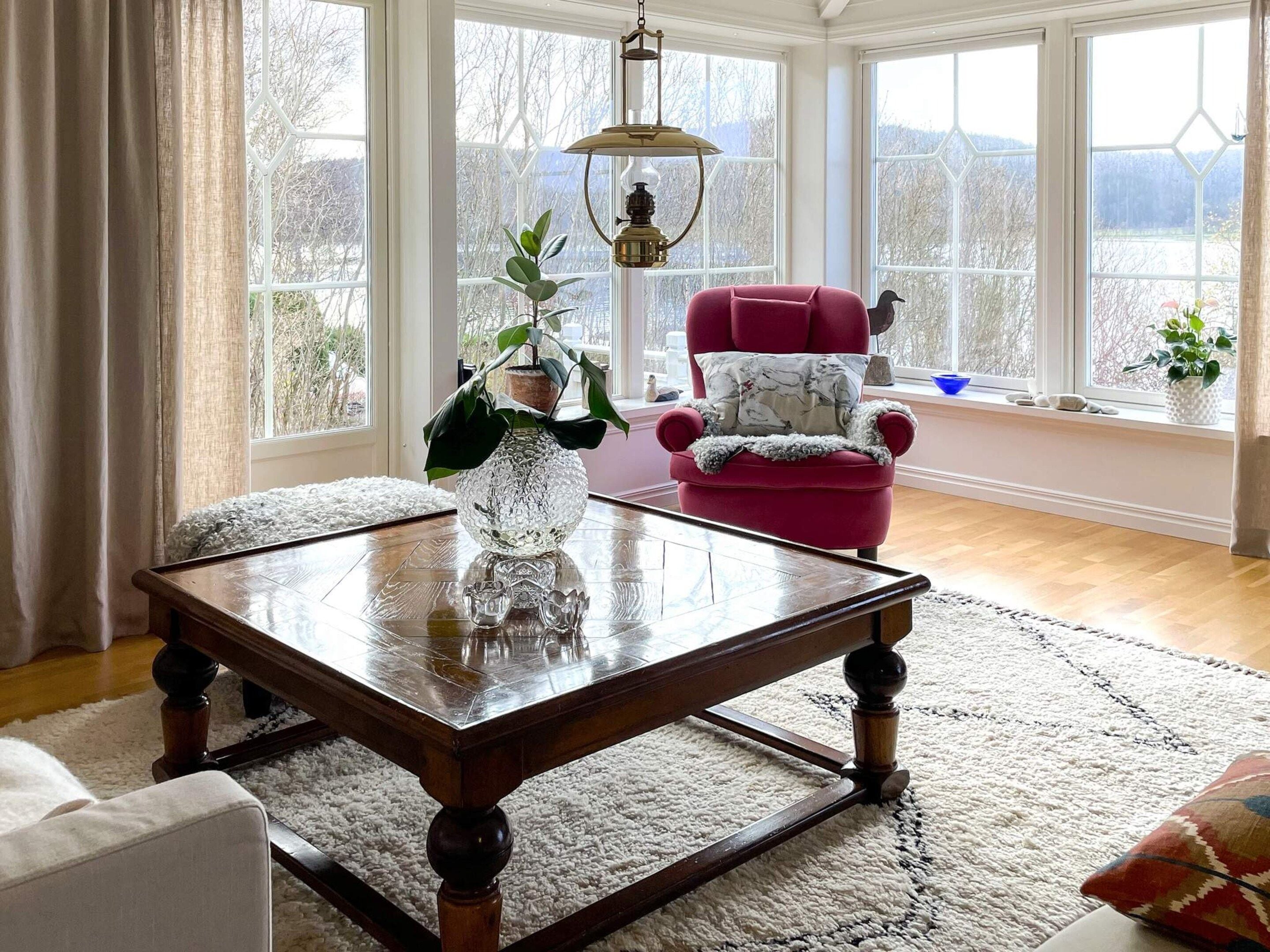 Quarto com mesa de centro de madeira, uma cadeira vermelha, uma poltrona branca e uma janela grande com vista para árvores e montanhas.