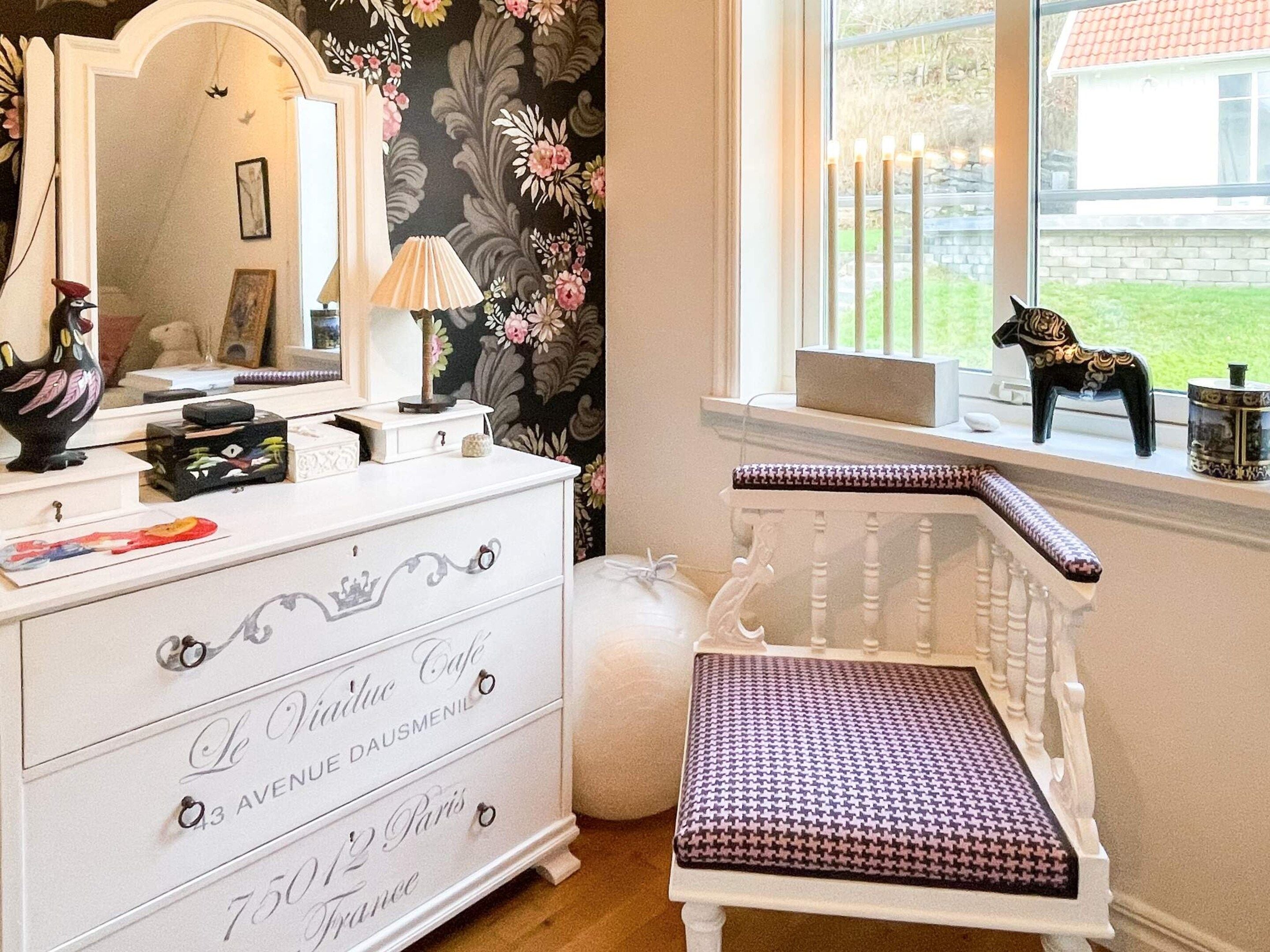Een kamer met een witte ladekast, een spiegel, een lamp en een raam met uitzicht op groen.
