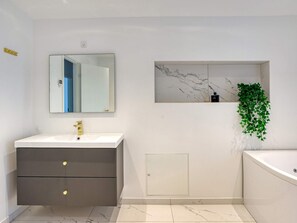 A modern bathroom with a white vanity, a large mirror, a marble backsplash, a green plant, and a bathtub.