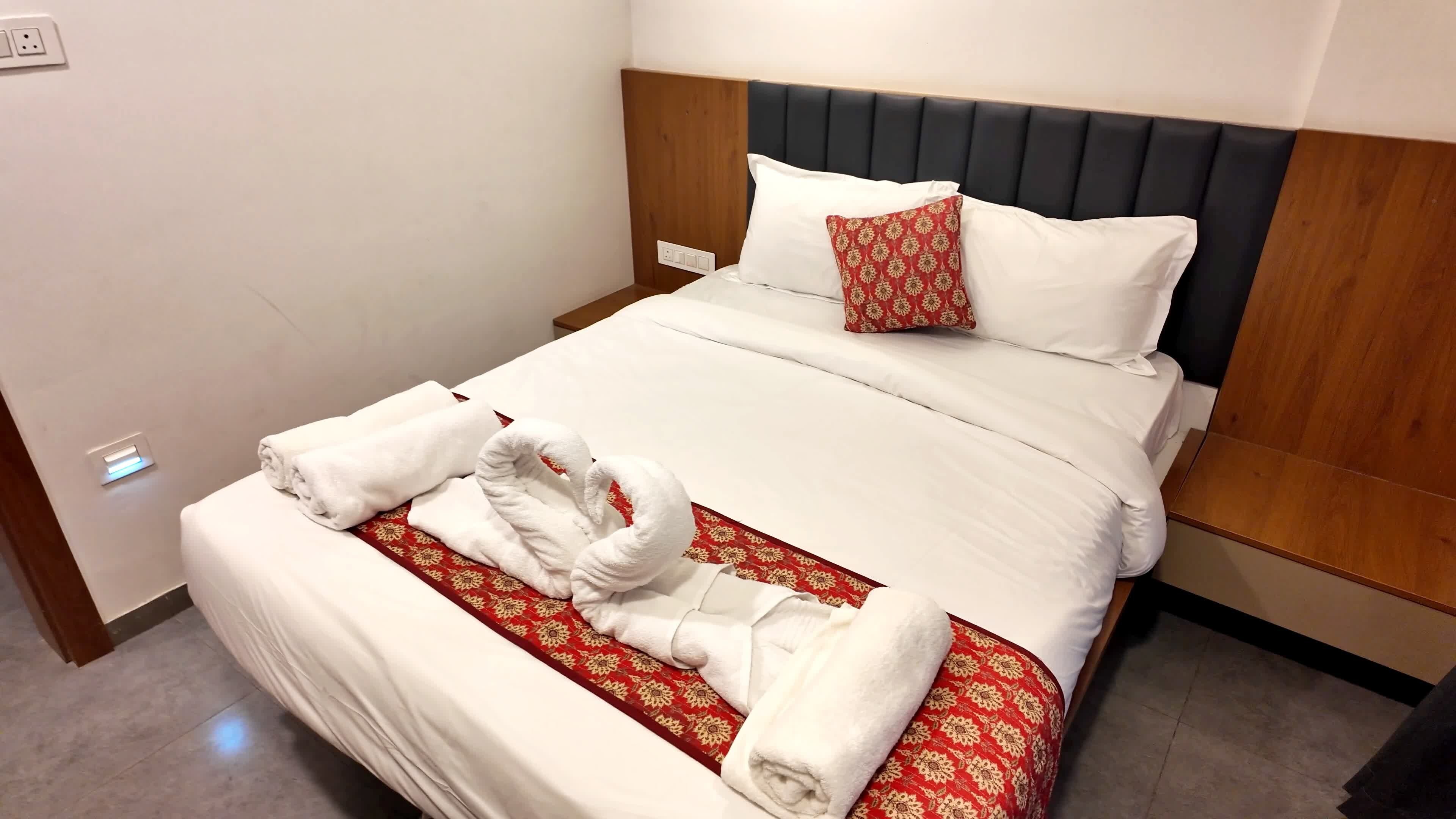 A hotel room with a neatly made bed, a wooden headboard, and a red patterned bedspread.