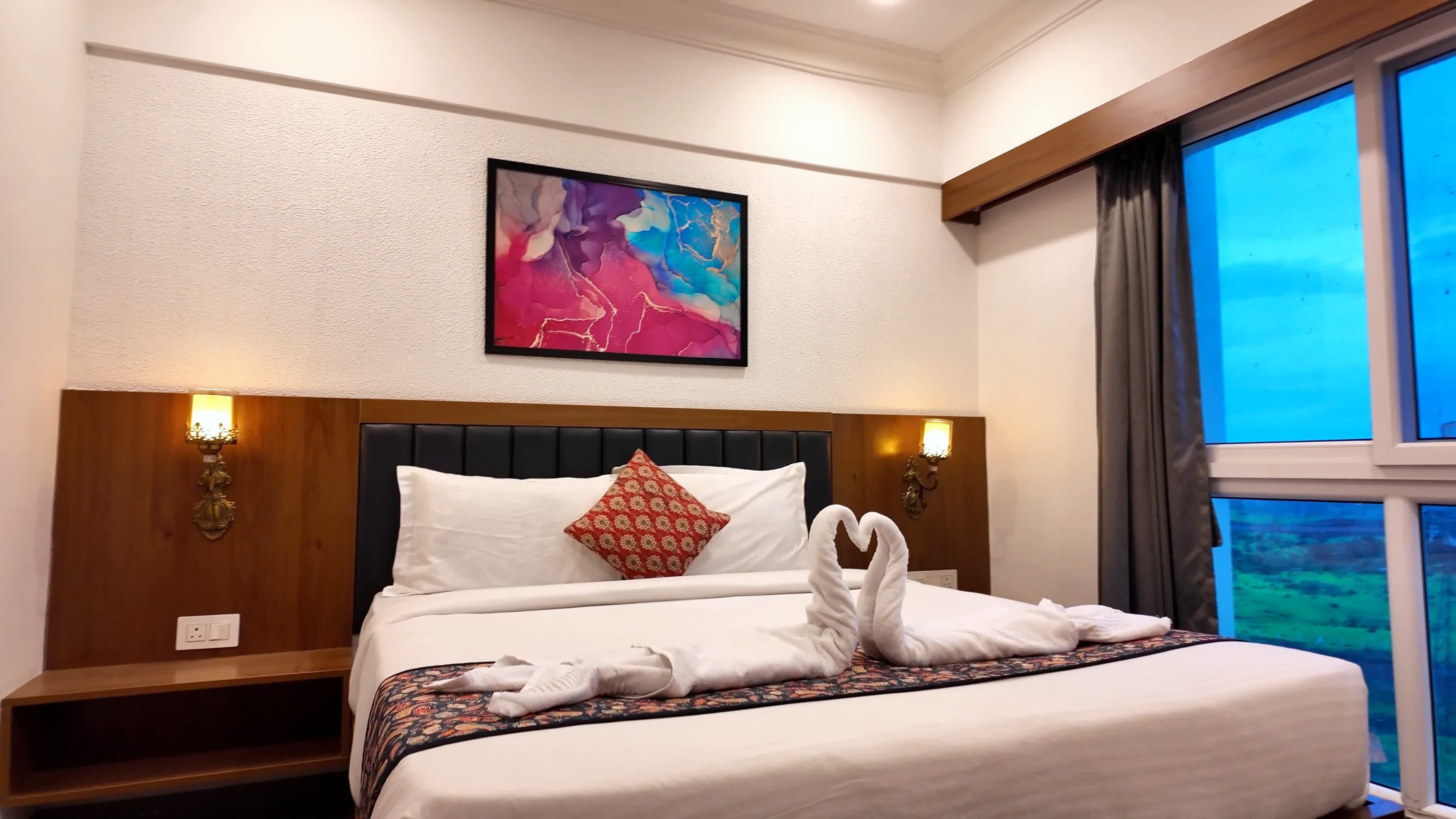 A hotel room with a bed, a painting, a nightstand, and a window with a view.