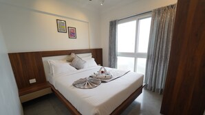 Een hotelkamer met een groot bed, een houten hoofdeinde, twee aan de muur bevestigde prenten, een raam met gordijnen en een houten deur.
