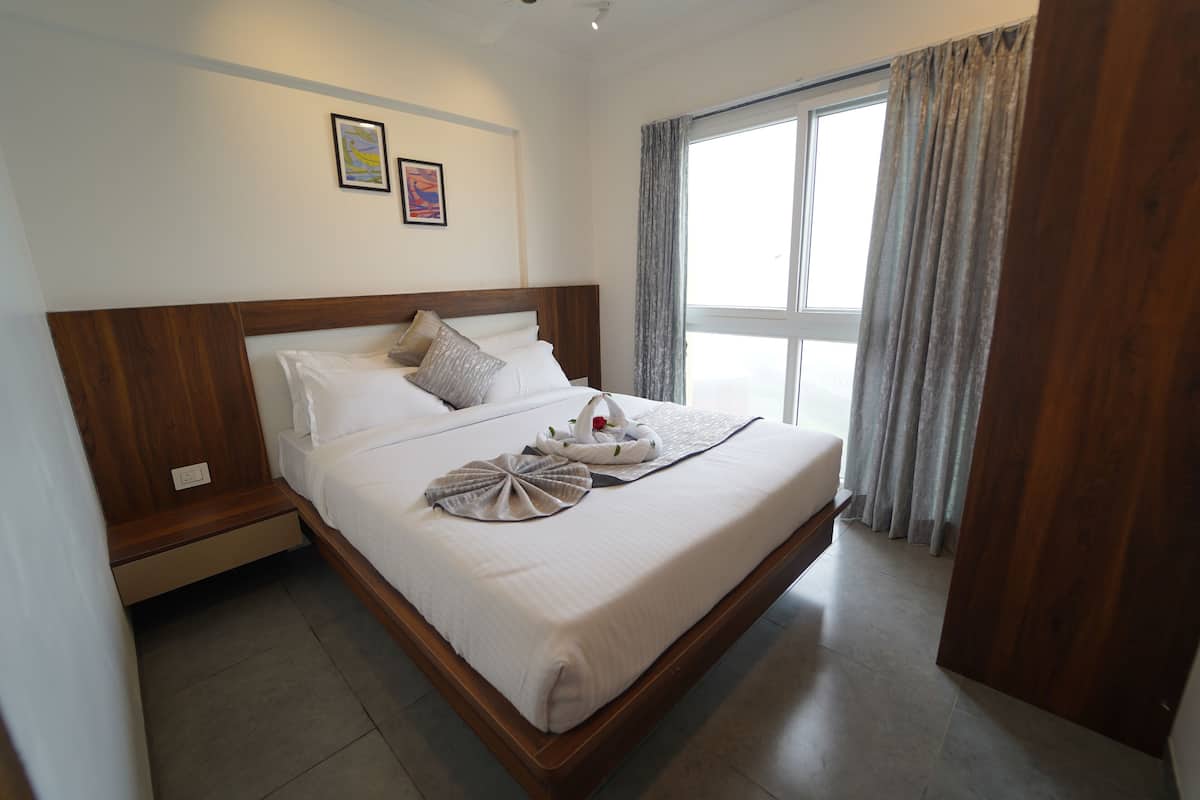 Habitación de hotel con una cama grande, cabecero de madera, dos cuadros colgados en la pared, una ventana con cortinas y una puerta de madera.