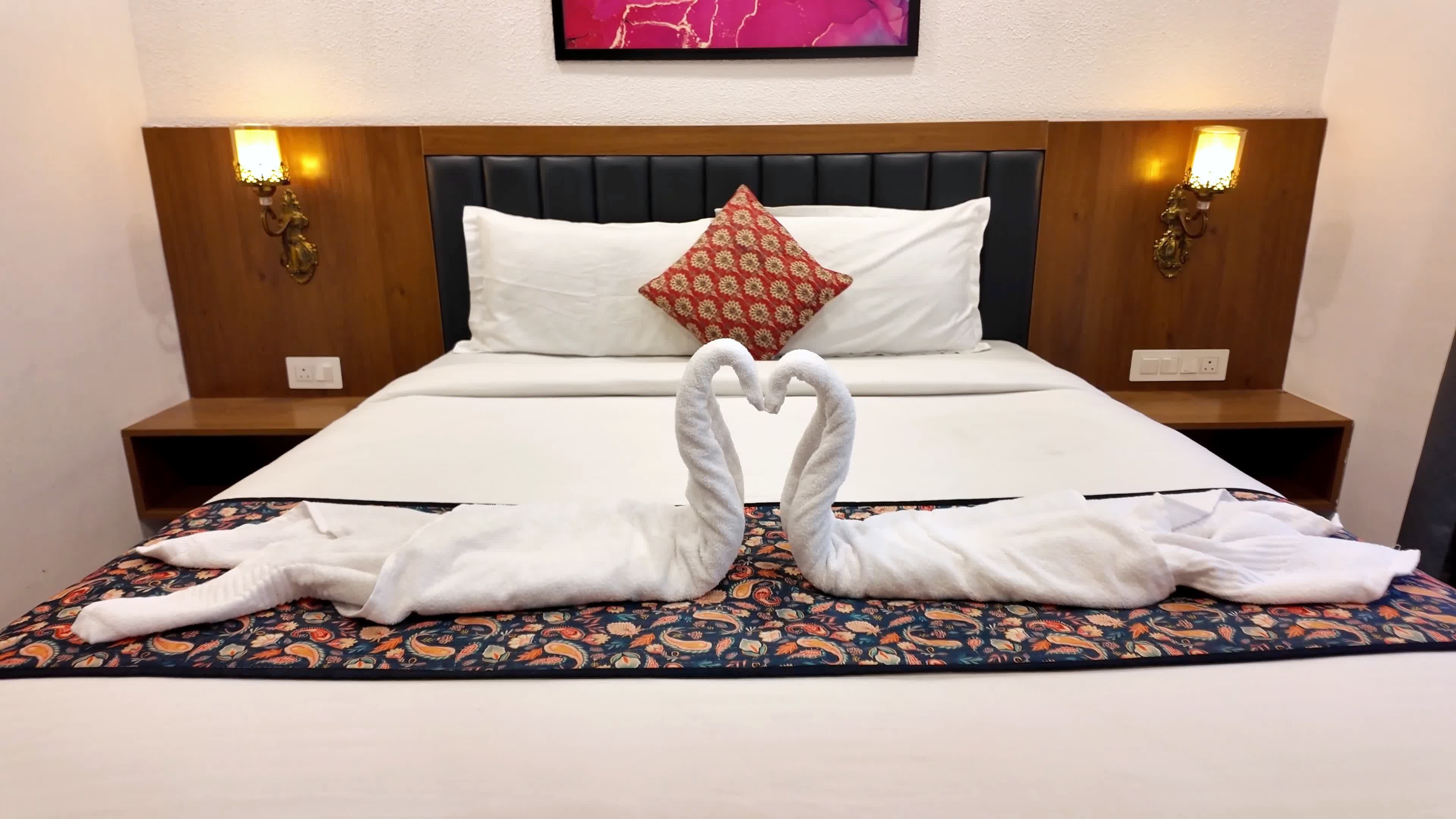 Quarto de hotel com cama, duas mesas de cabeceira, abajur fixado na parede e almofada decorativa.