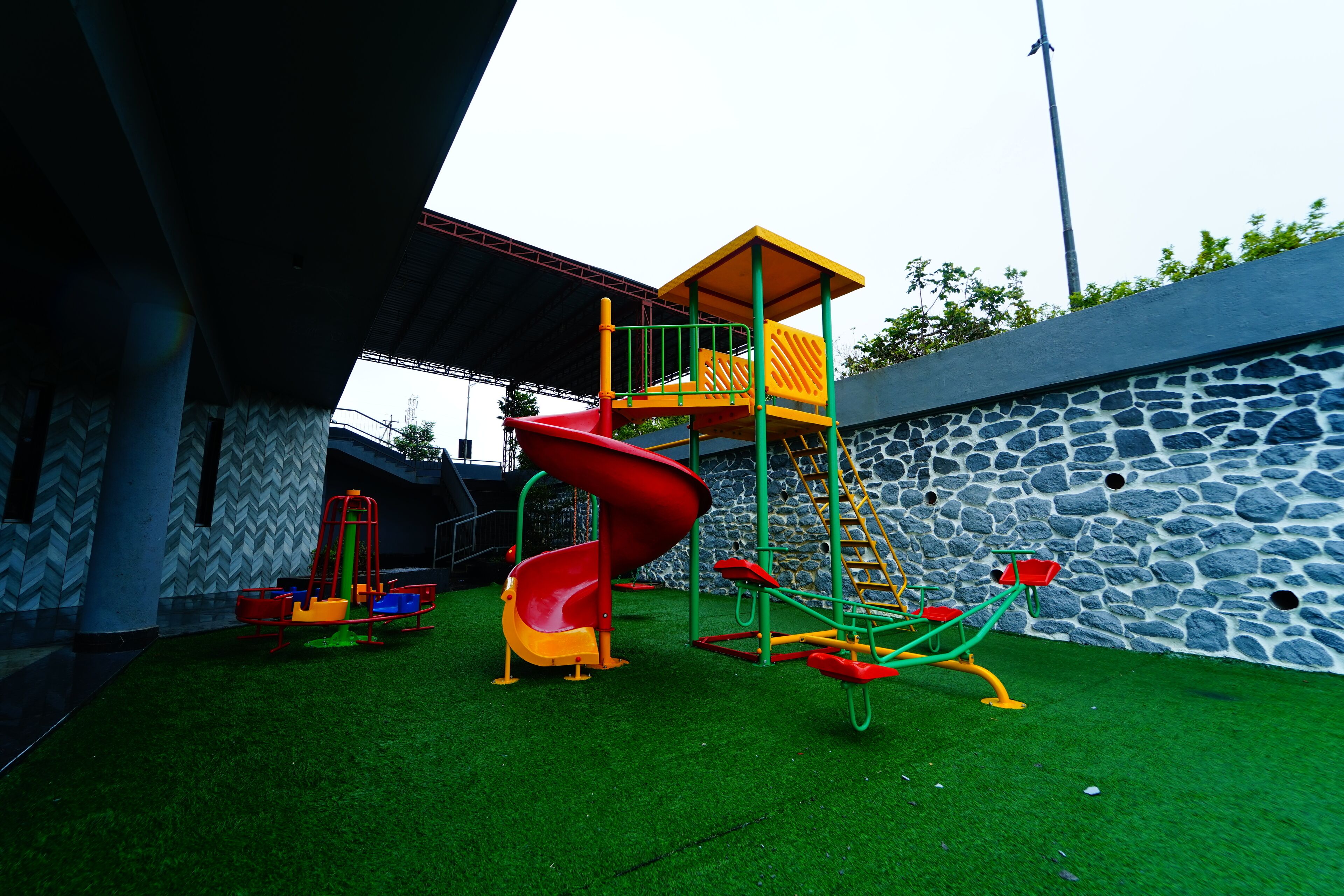 A playground with a red slide and climbing structures, surrounded by a stone wall and artificial grass.