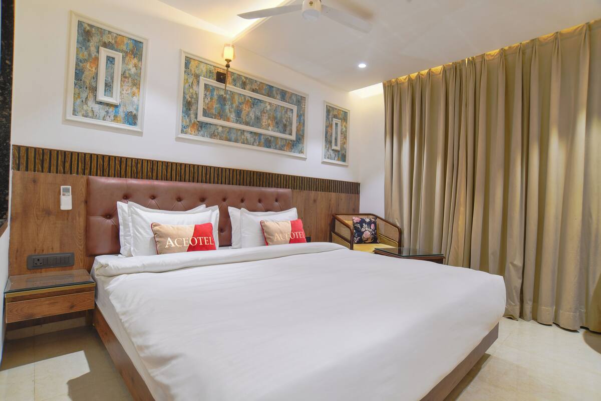 A hotel room with a large bed, a headboard, a ceiling fan, artwork on the wall, and a window with curtains.