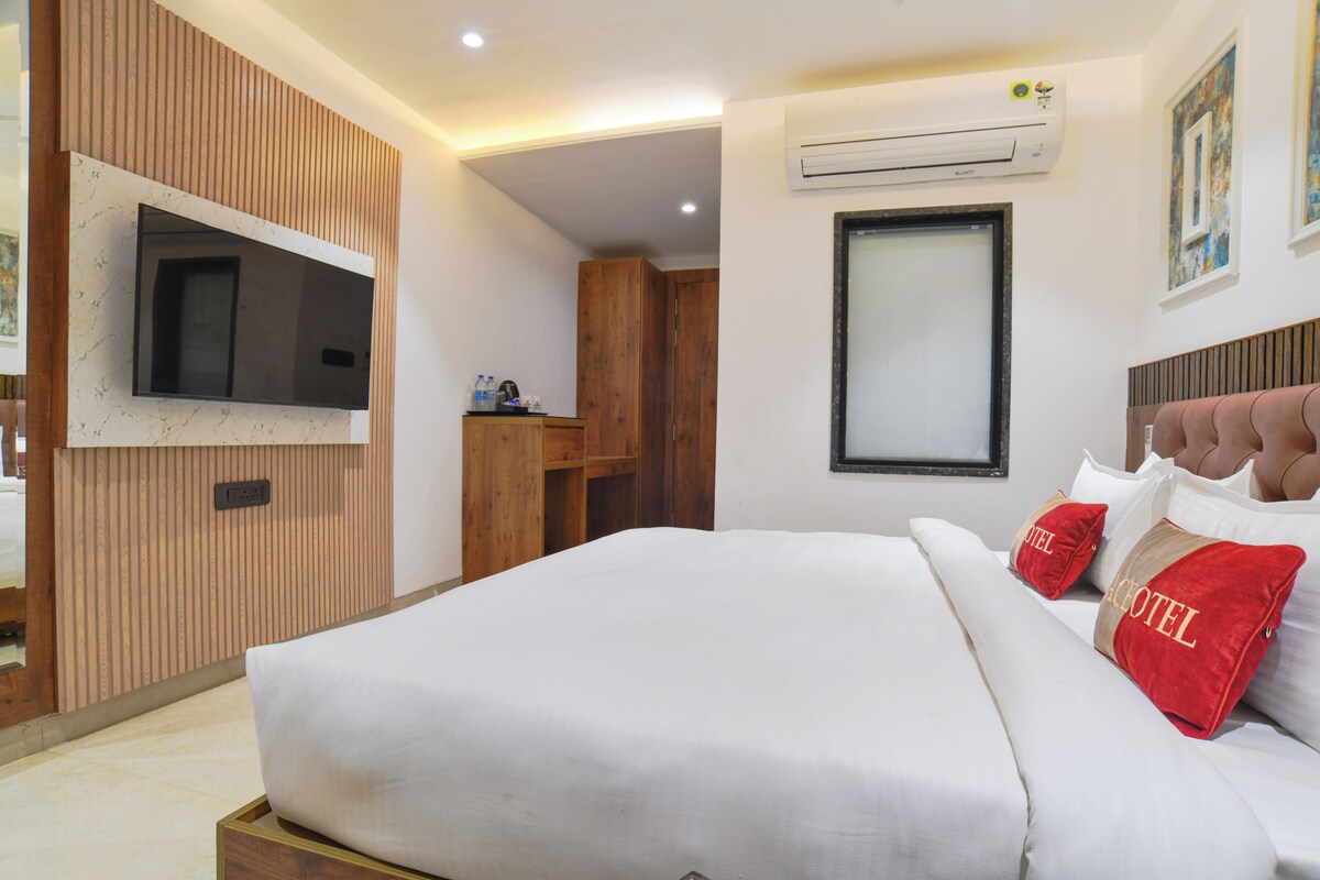 A hotel room with a bed, a TV mounted on the wall, an air conditioning unit, and a wooden wardrobe.