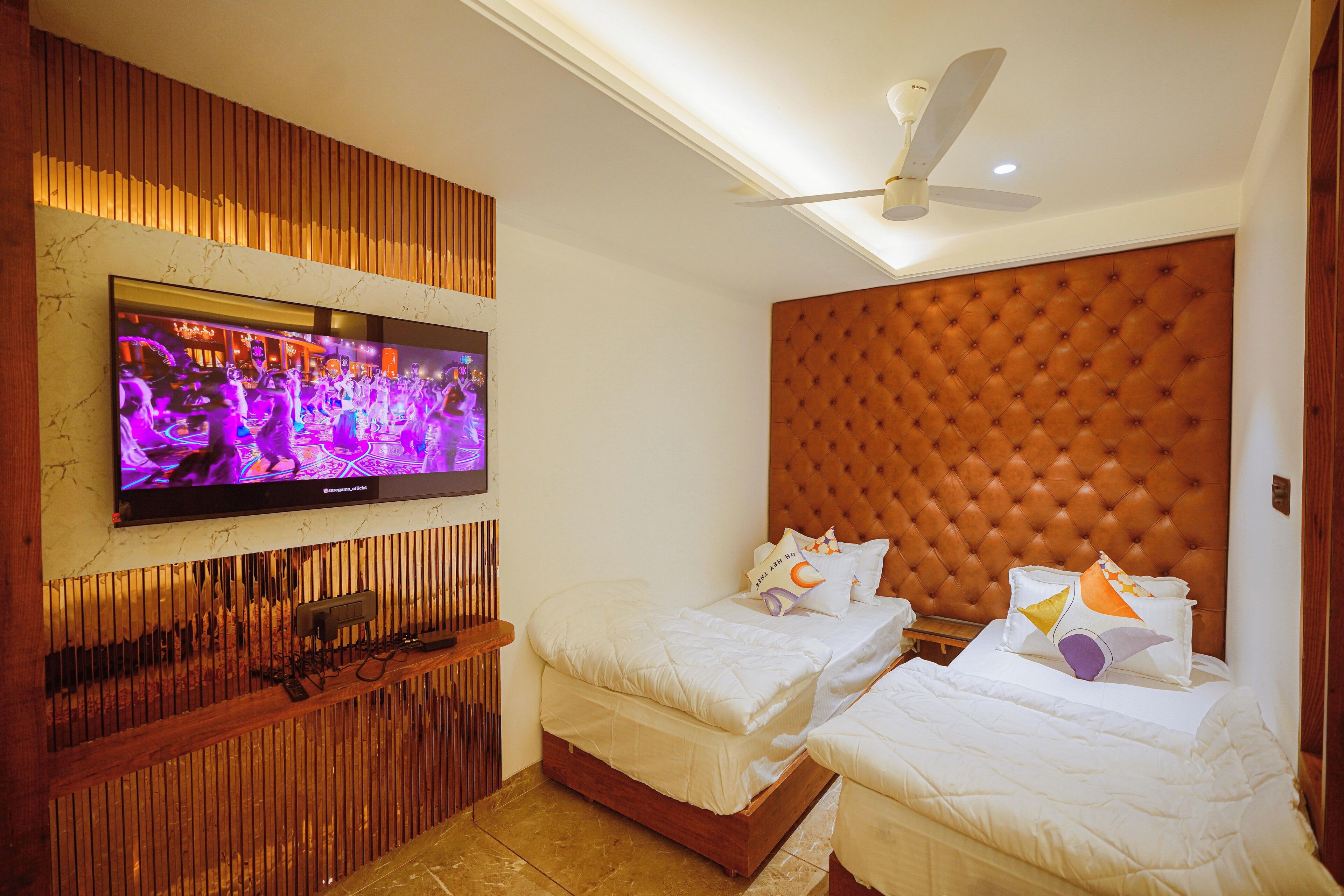 A hotel room with a large flat-screen TV mounted on the wall, a tufted headboard, two beds with white bedding, and a ceiling fan.