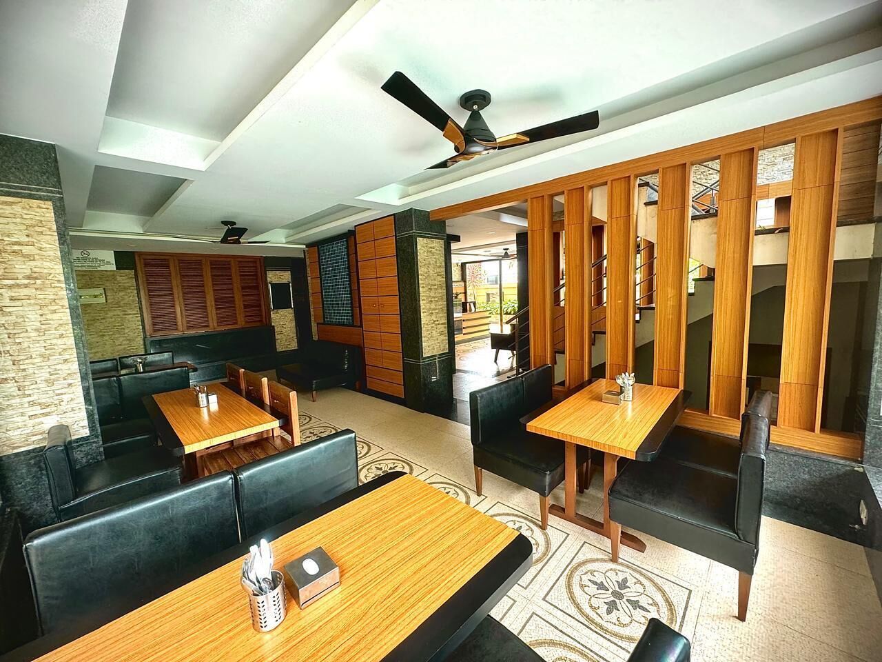 Un restaurante con mesas de madera, asientos tapizados en negro y un ventilador de techo, con vistas al exterior.
