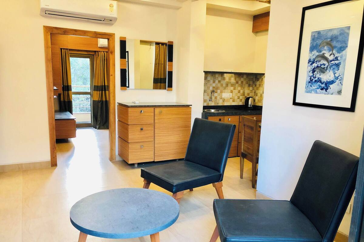 A modern living room with two black chairs and a round table, a wooden cabinet, a kitchen area with a tiled backsplash, and a framed artwork on the wall.