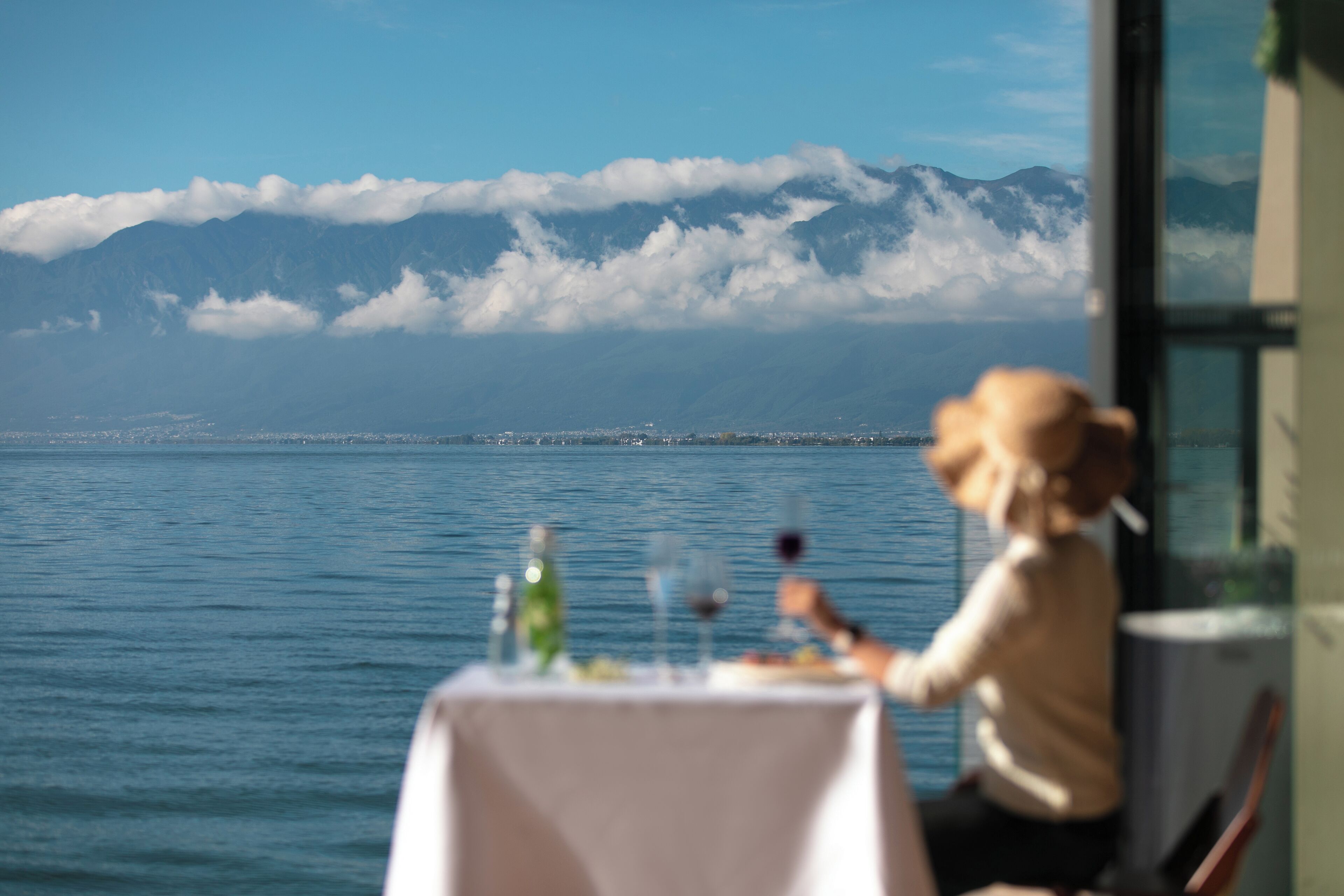 A table with a bottle and glasses, a person sitting at the table, and a view of mountains and water.