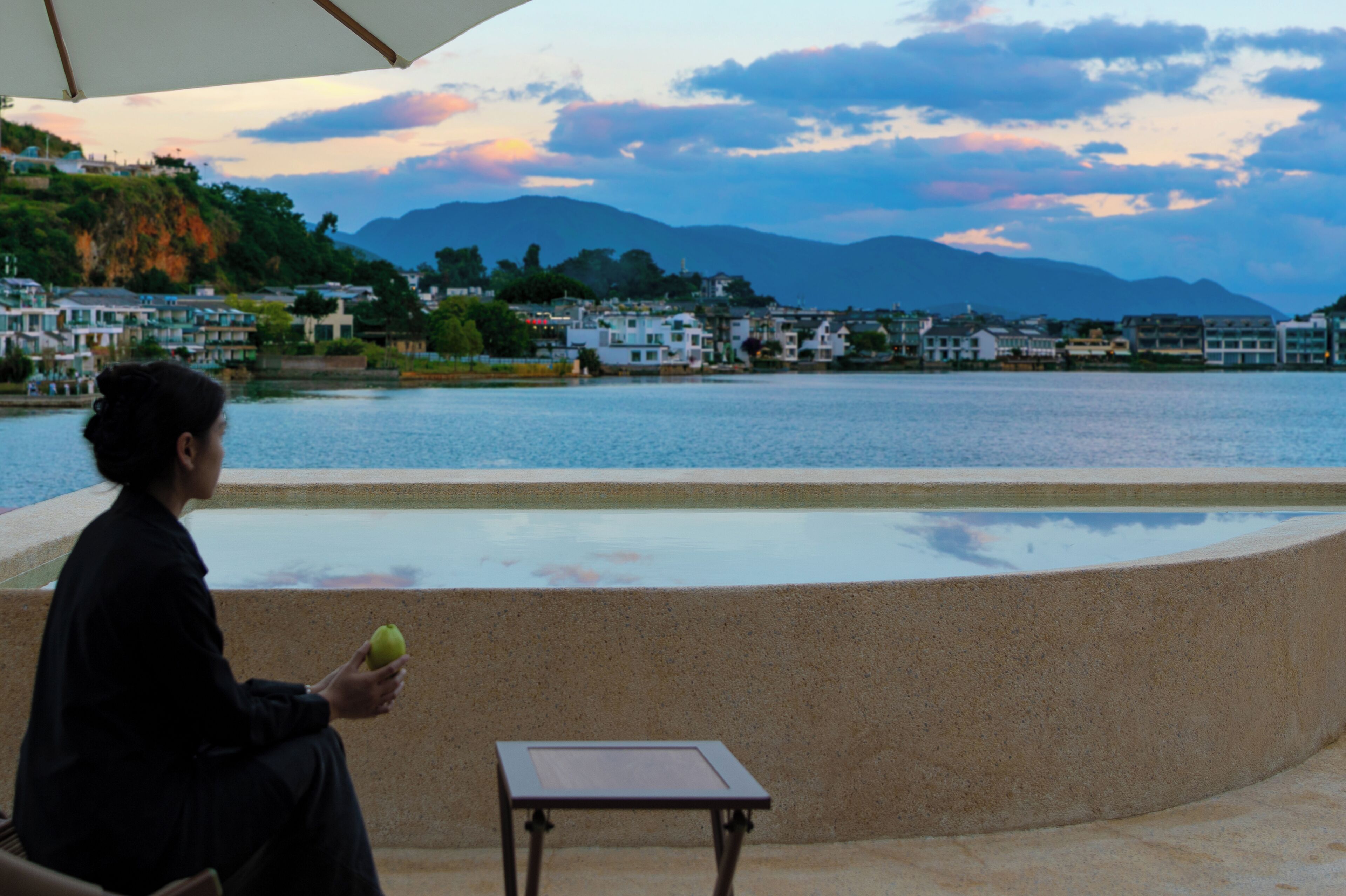 A person sitting by a waterfront with a table and umbrella, overlooking a town and mountains.