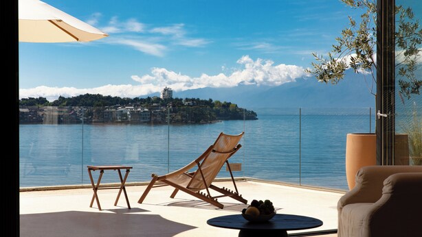 A balcony with a view of a lake, a wooden chair, a small table, and a large potted plant.